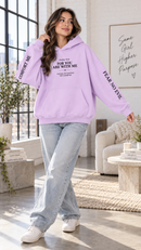 A woman modeling a light purple hoodie with printed scripture text on the front and 'COMFORT ME' and 'FEAR NO Evil' on the sleeves, paired with light-wash jeans and white sneakers in a bright, modern living room.